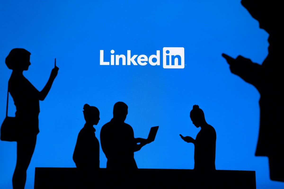 Several silhouettes of people interact with devices in front of a large LinkedIn logo displayed on a blue background