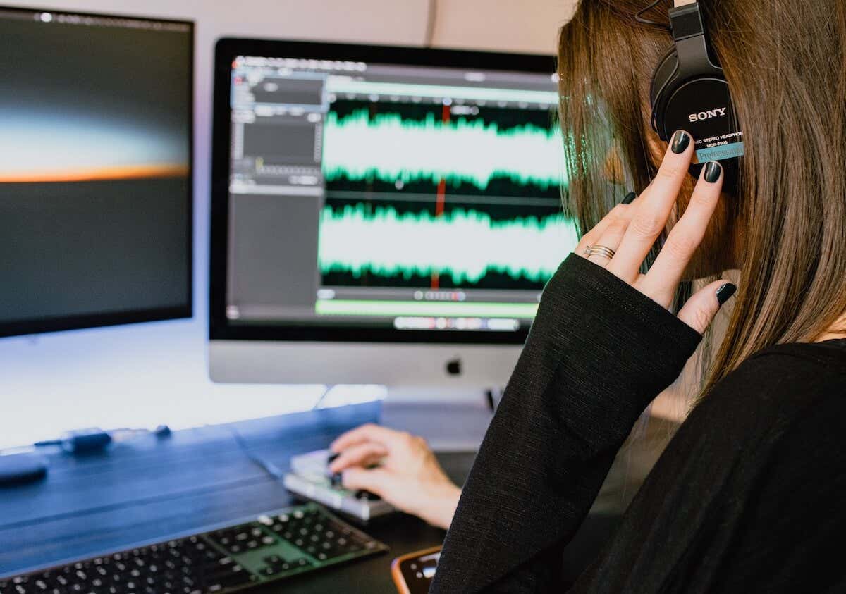 A woman adjusts headphones while working at a computer displaying audio waveforms