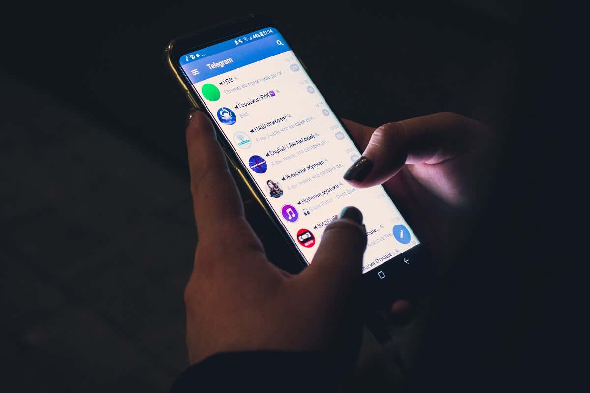 A person's hands interact with a smartphone displaying the Telegram app, actively scrolling through a list of channels in a dimly lit environment