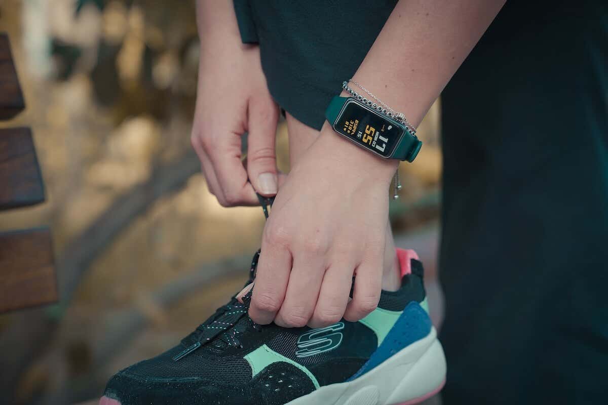 A person ties a shoelace on a black and teal sneaker while wearing a dark green smartwatch displaying "11 55" and other data