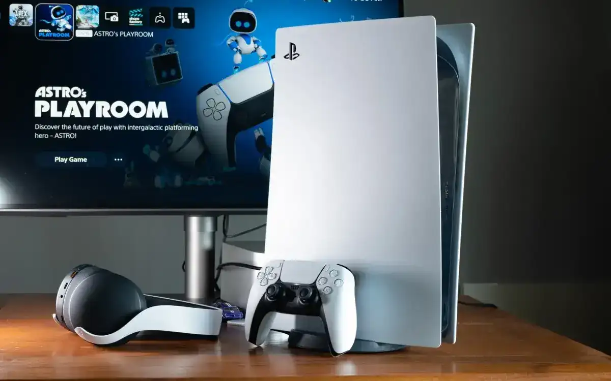A white PlayStation 5 console rests on a wooden desk, next to a controller and headset; a monitor displays the Astro's Playroom game